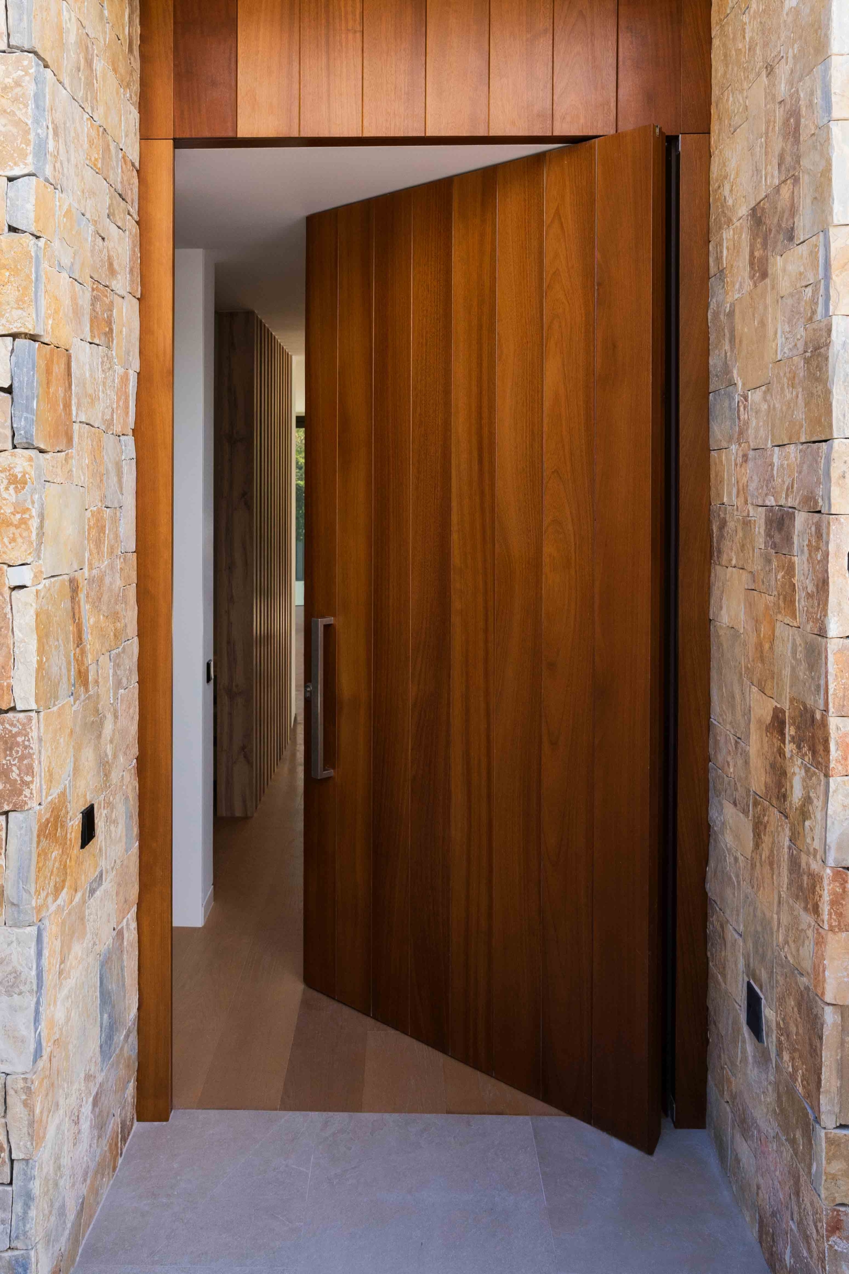 Puerta pivotante de madera dando acceso a interior de la vivienda contrastando con la fachada de piedra natural